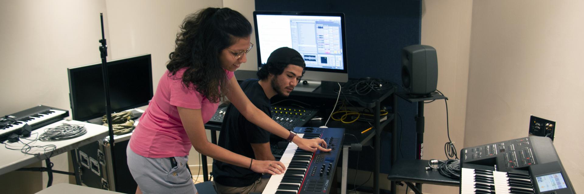 A female student plays the keyboard while a male student watches to record her in a studio.