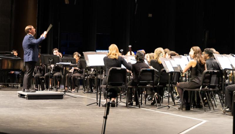 Concert band performs on stage with conductor, Benjamin Diden